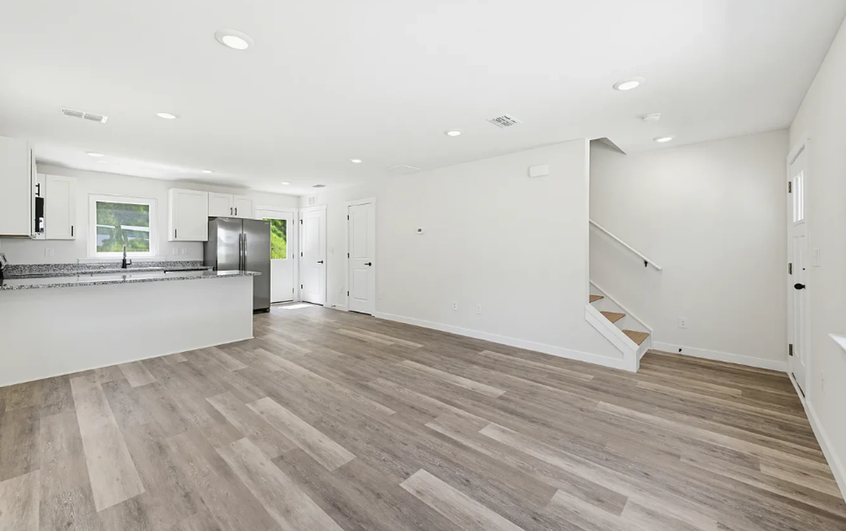 Open living area in a Legacy Signature Rentals home in Conover, NC — carpet-free plank flooring, white walls, a granite breakfast bar with stainless-steel refrigerator behind it and a staircase to the right