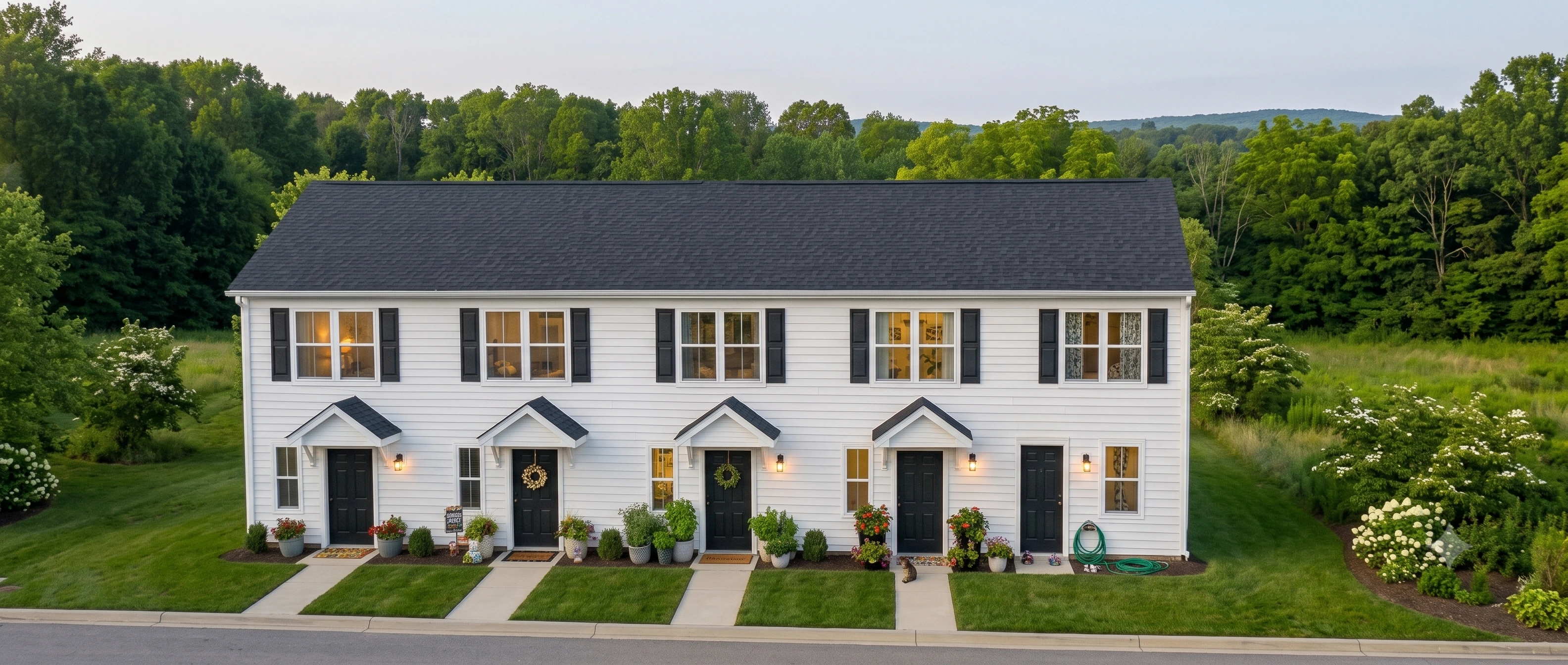 Row of four attached Legacy Signature Rentals townhomes for rent in Newton, NC at dusk — white siding with black shutters, gabled porch roofs over black front doors, lit windows and a private walkway to each entry
