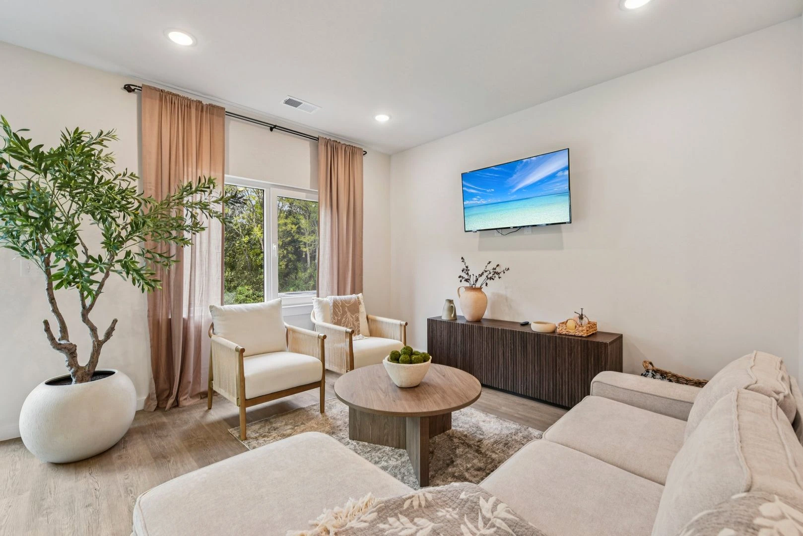Living room in a Legacy Signature Rentals home in Newton, NC — carpet-free plank flooring, a cream sofa and two armchairs around a round wooden coffee table, a wall-mounted TV above a dark ribbed sideboard and an olive tree by the window