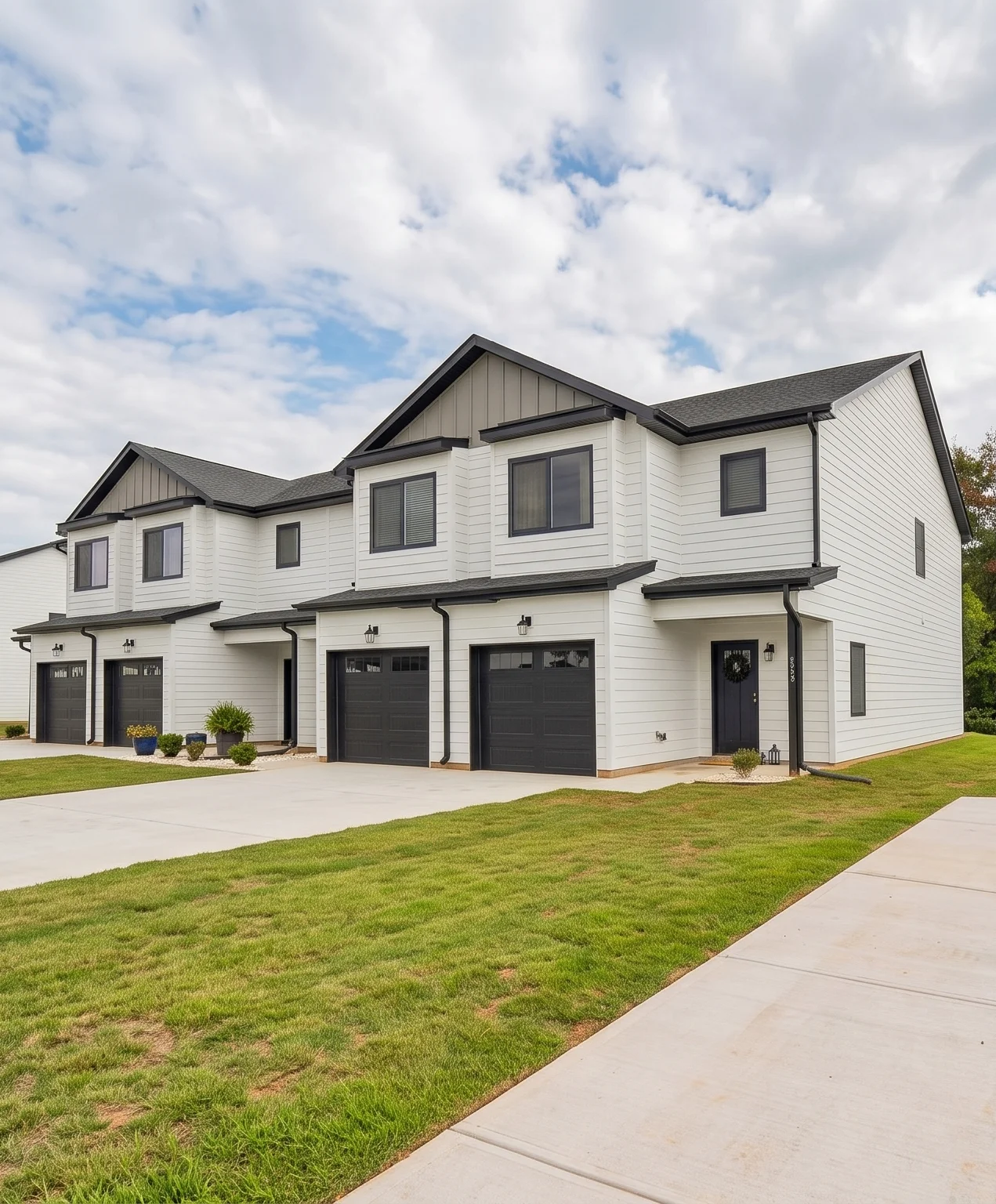 Legacy Signature Rentals townhomes for rent in Newton, NC seen from the driveway — white siding with dark grey board-and-batten gables, black garage doors and porch lanterns, and a mown lawn along the walkway
